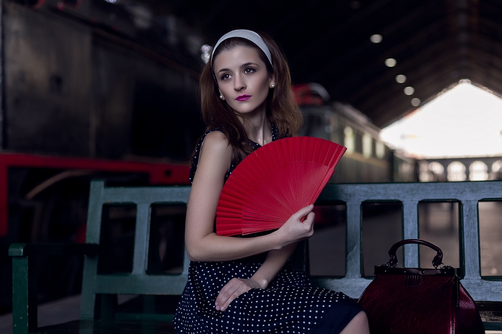 Chica sentada en un banco en una estación de tren con un abanico rojo.