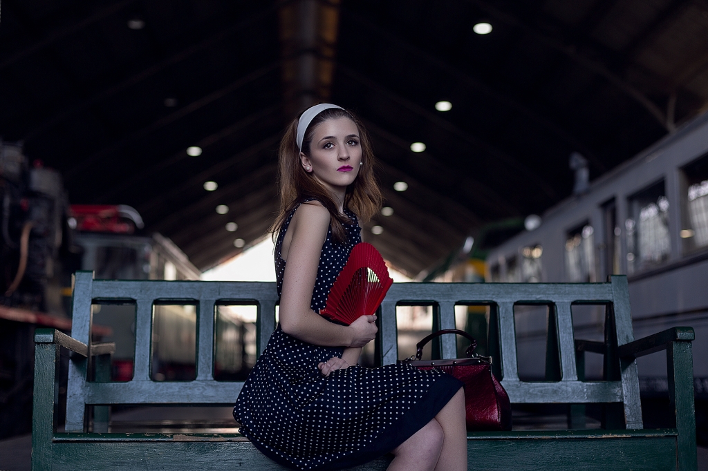 Chica sentada en un banco en una estación de tren con un abanico rojo y los trenes a ambos lados.