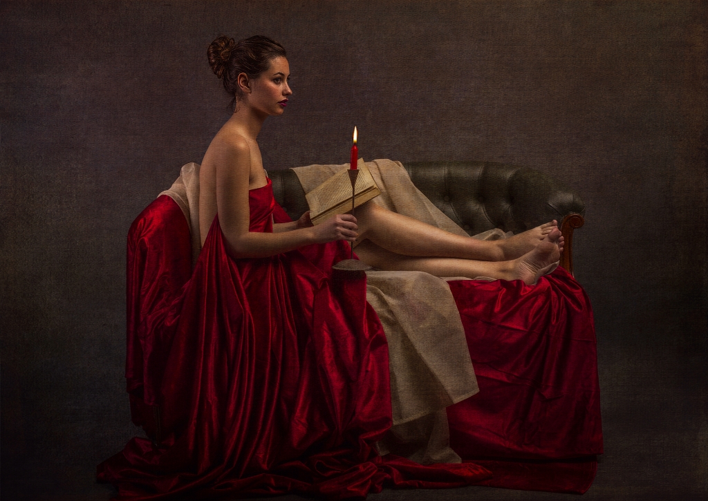 Mujer vestida de rojo tumbada en un sofá con una vela y un libro en la mano.