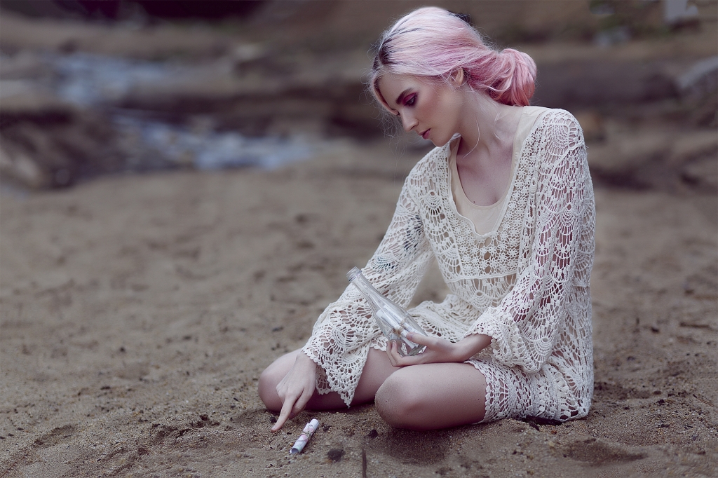 Chica con el pelo rosa sentada en la arena con una botella llena de mensajes.