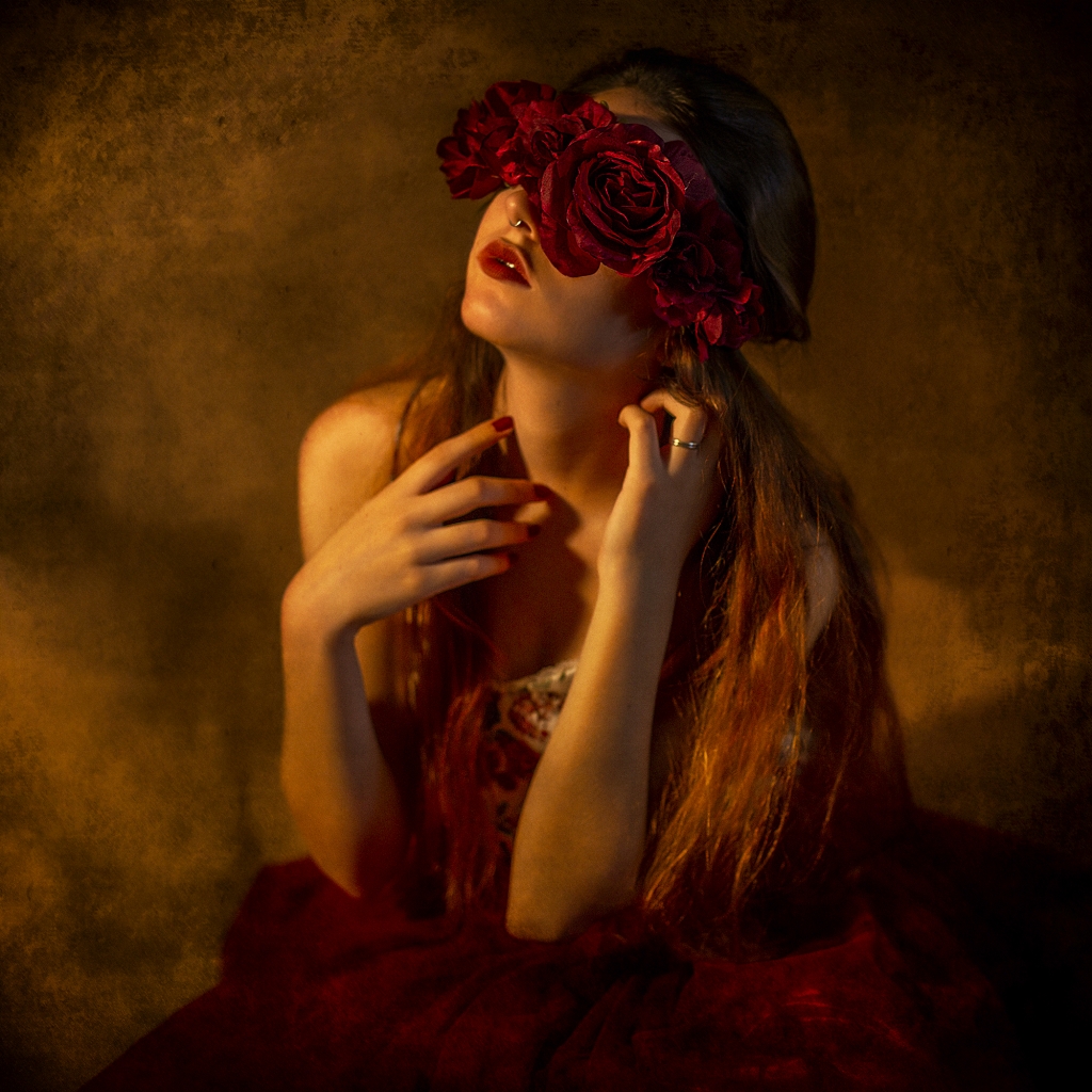Mujer sentada con falda roja, brazos cruzados en el pecho y flores cubriéndole los ojos.
