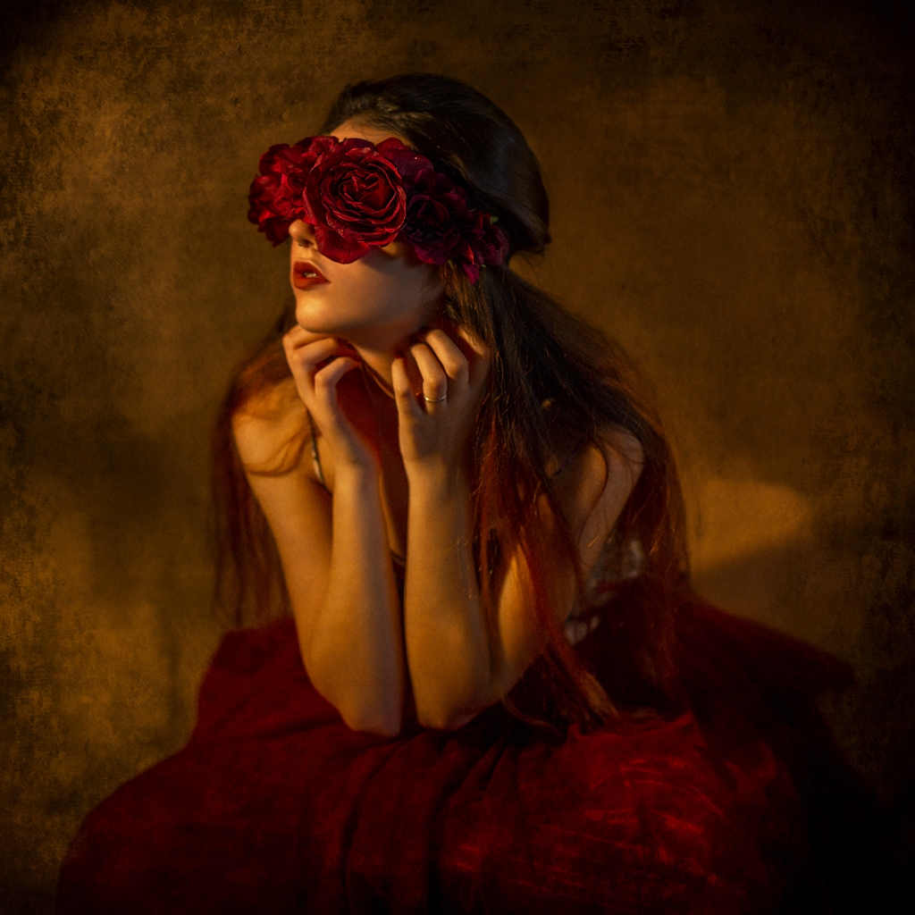 Mujer sentada con falda roja, brazos cruzados en el pecho y flores cubriéndole los ojos.