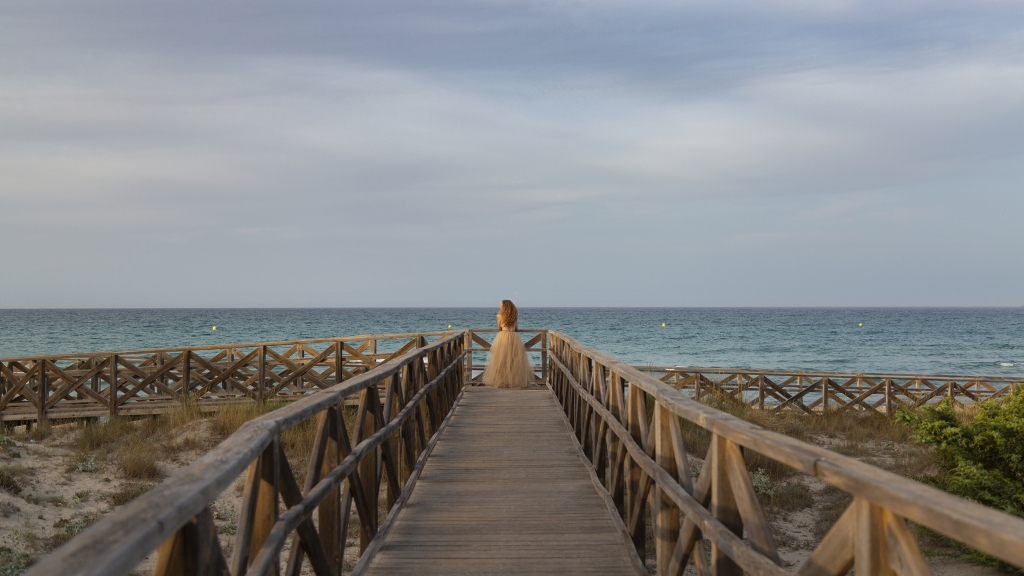 Mujer al final de una pasarela de madera, mirando al mar.