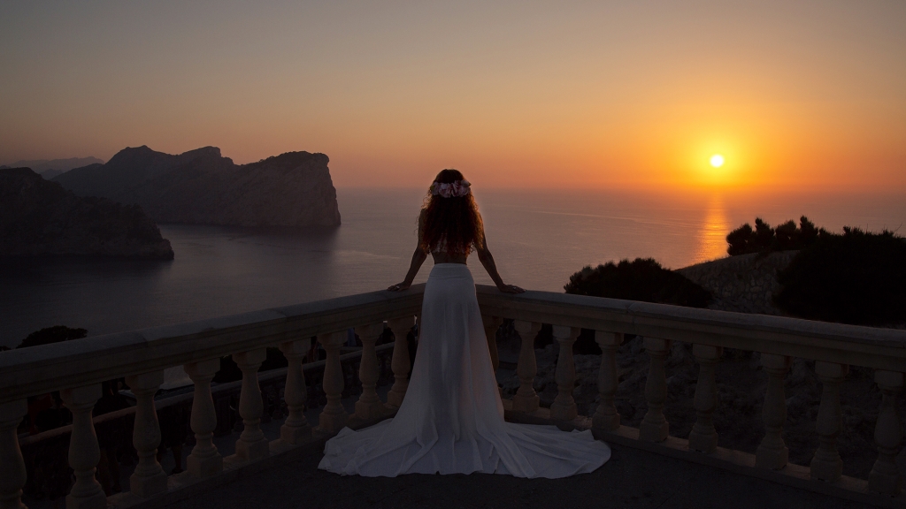 Mujer vestida de blanco y apoyada en una barandilla mirando la puesta de sol.