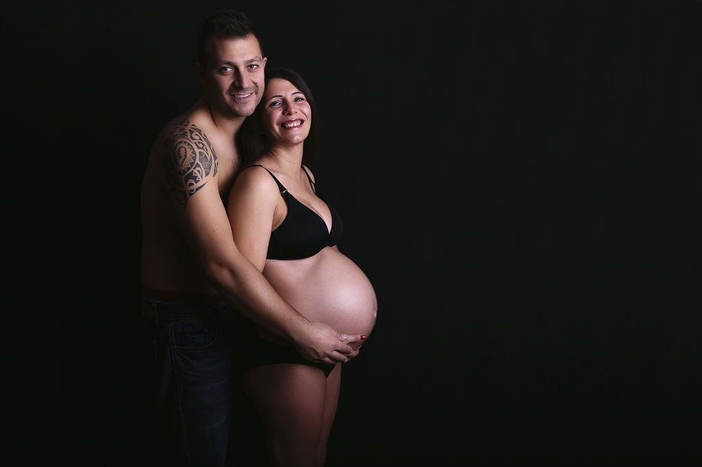 Mujer embarazada en estudio con fondo negro abrazada por su pareja.