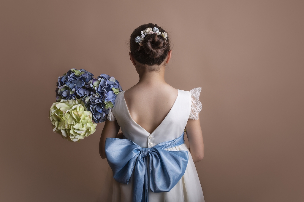 Niña vestida de Comunión con un gran lazo azul y un ramo de hortensias azules.